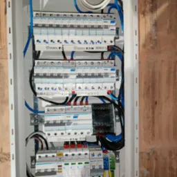 Rozdzielnica elektryczna z zainstalowanymi bezpiecznikami i przewodami w kolorach niebieskim, czarnym, żółto-zielonym i białym, zamontowana na ścianie z cegły.