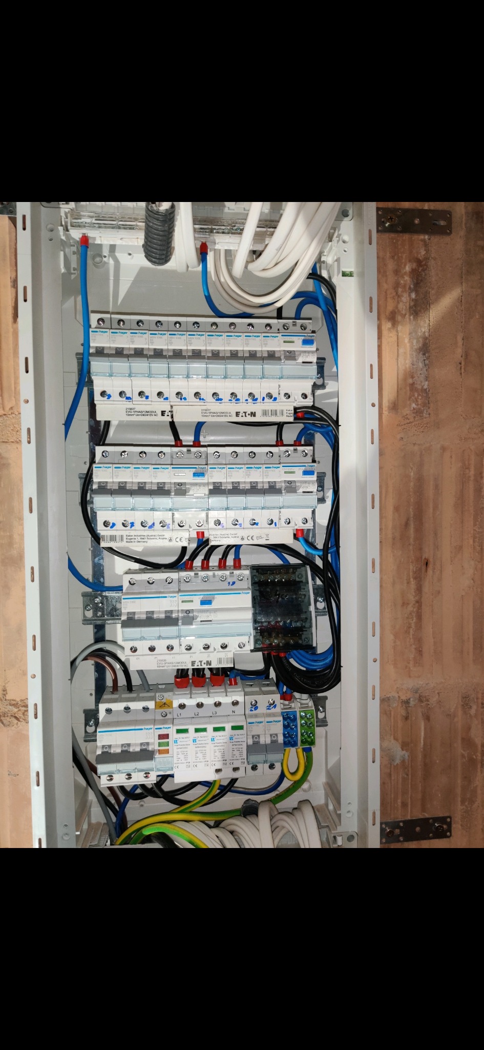 Rozdzielnica elektryczna z zainstalowanymi bezpiecznikami i przewodami w kolorach niebieskim, czarnym, żółto-zielonym i białym, zamontowana na ścianie z cegły.