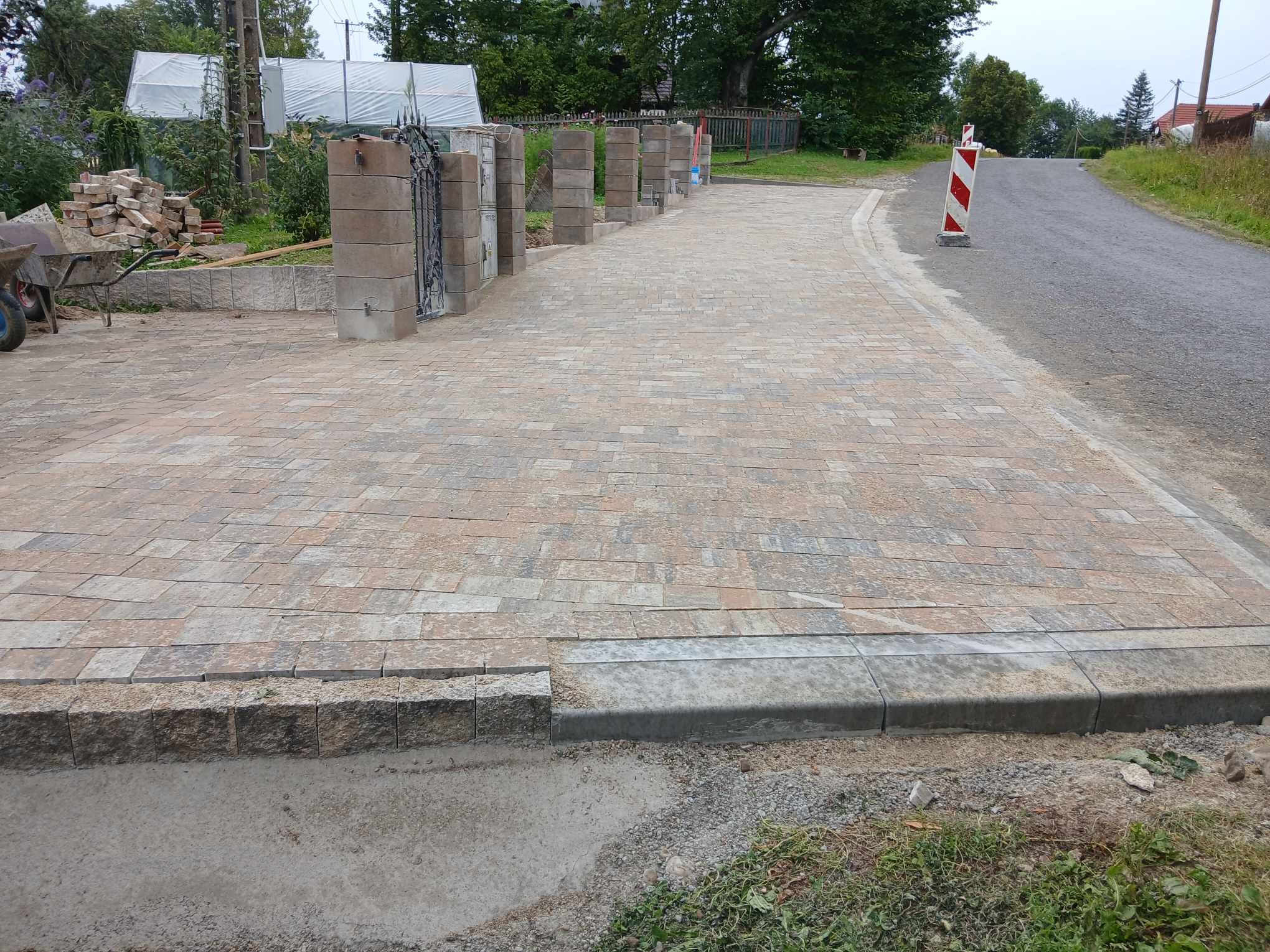 Ułożona kostka brukowa w odcieniach beżu i szarości, z widocznymi obrzeżami betonowymi i słupkami z bloków betonowych przy wjeździe, w tle zieleń drzew i krzewów.