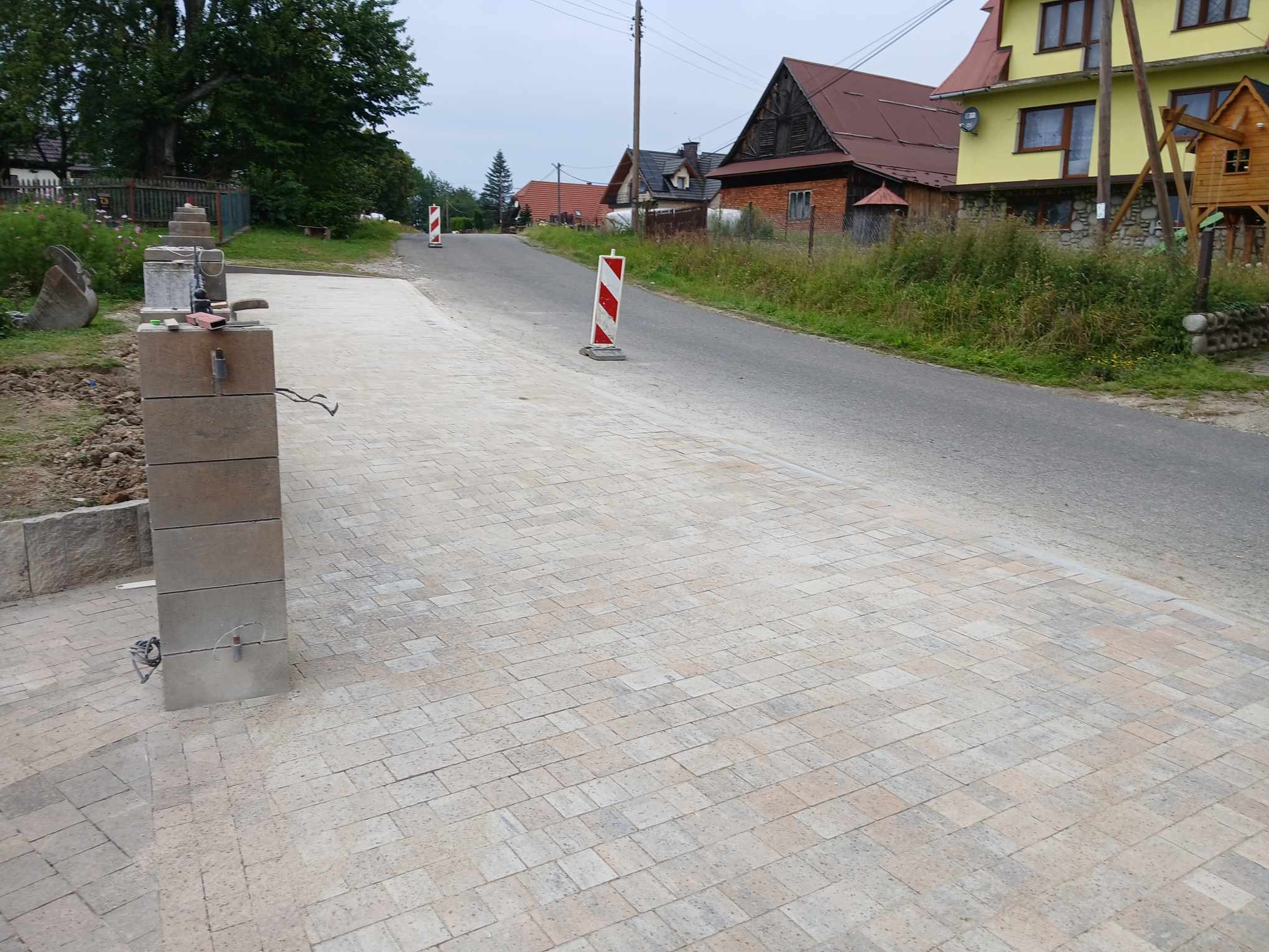 Nowo ułożona, beżowo-szara kostka brukowa prowadząca do asfaltowej drogi w wiejskiej scenerii. Widoczne słupki z kostki brukowej w trakcie montażu i znaki drogowe.