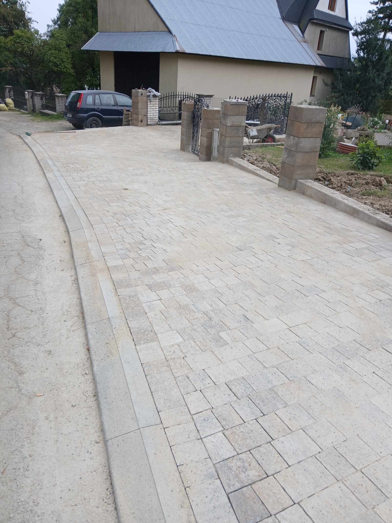 Szeroki podjazd z jasnej kostki brukowej prowadzący do domu z garażem, widoczne słupki ogrodzeniowe z szarego kamienia i czarny samochód osobowy w tle.