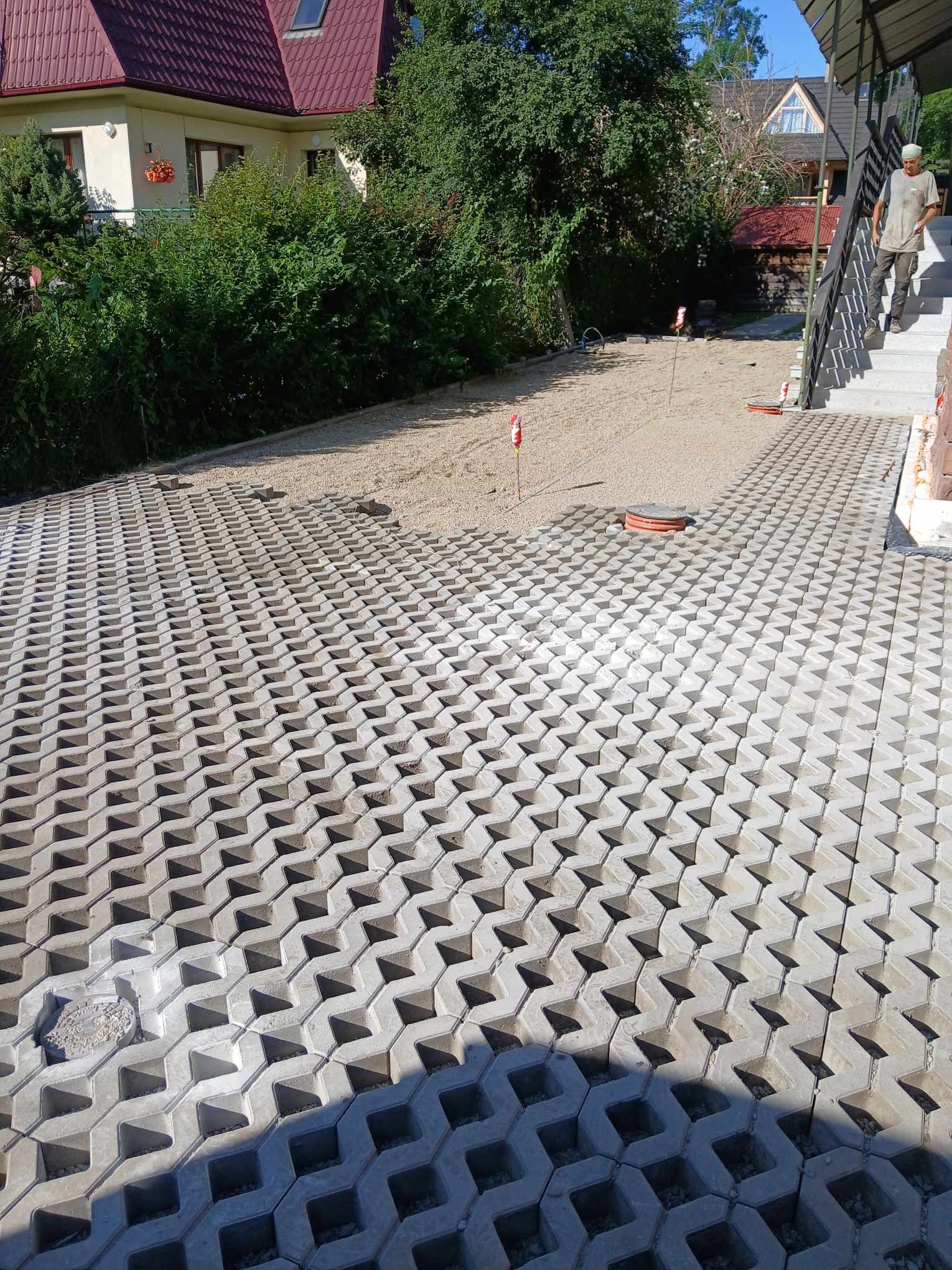 Ułożona ażurowa kostka brukowa na podjeździe, widoczny fragment utwardzonej drogi z kruszywa, mężczyzna schodzący po schodach w tle.