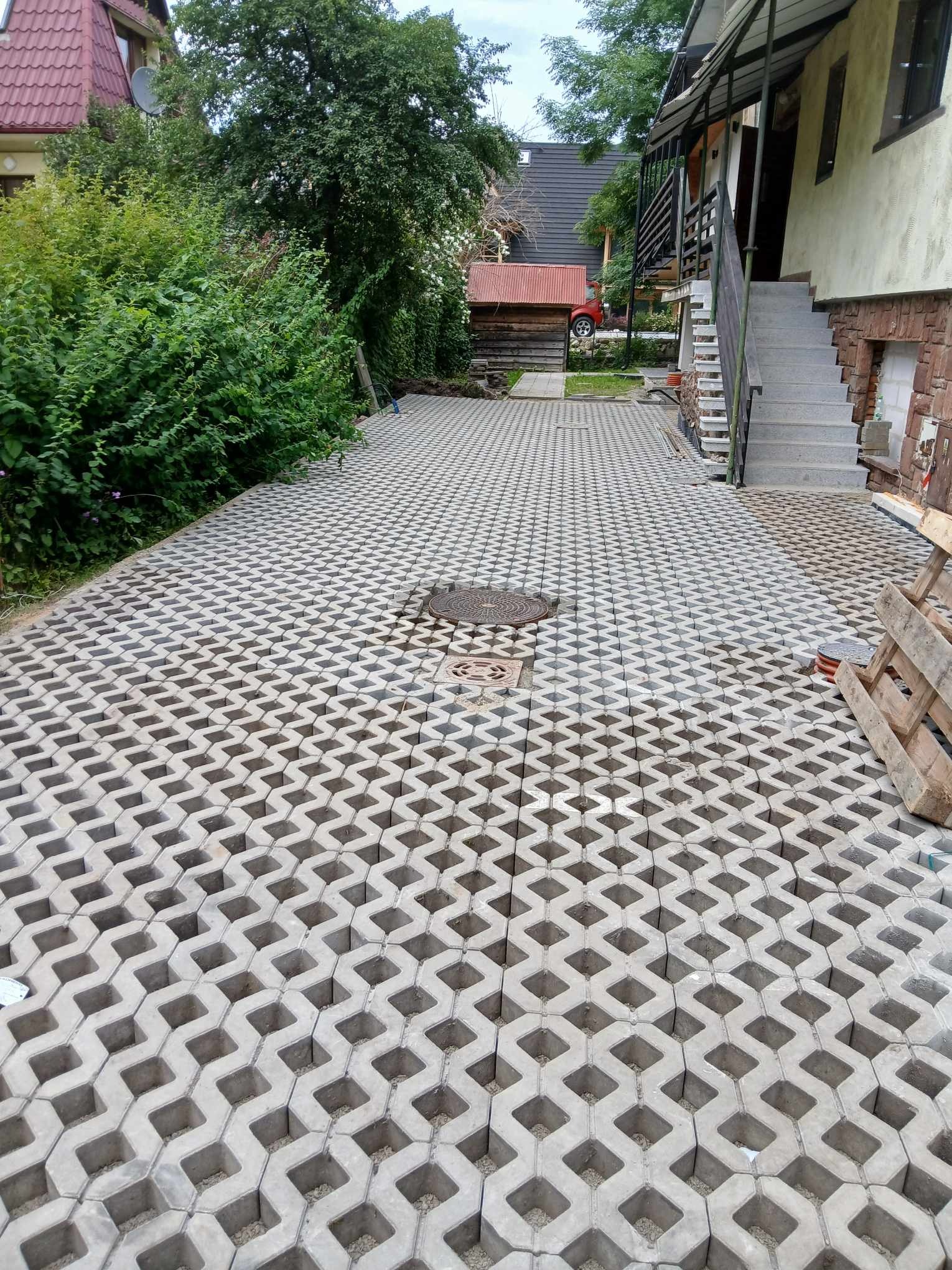 Szeroki, ukośny widok na szarą, ażurową kostkę brukową ułożoną na podjeździe, z widocznymi studzienkami kanalizacyjnymi, paletą z boku i zielenią w tle.