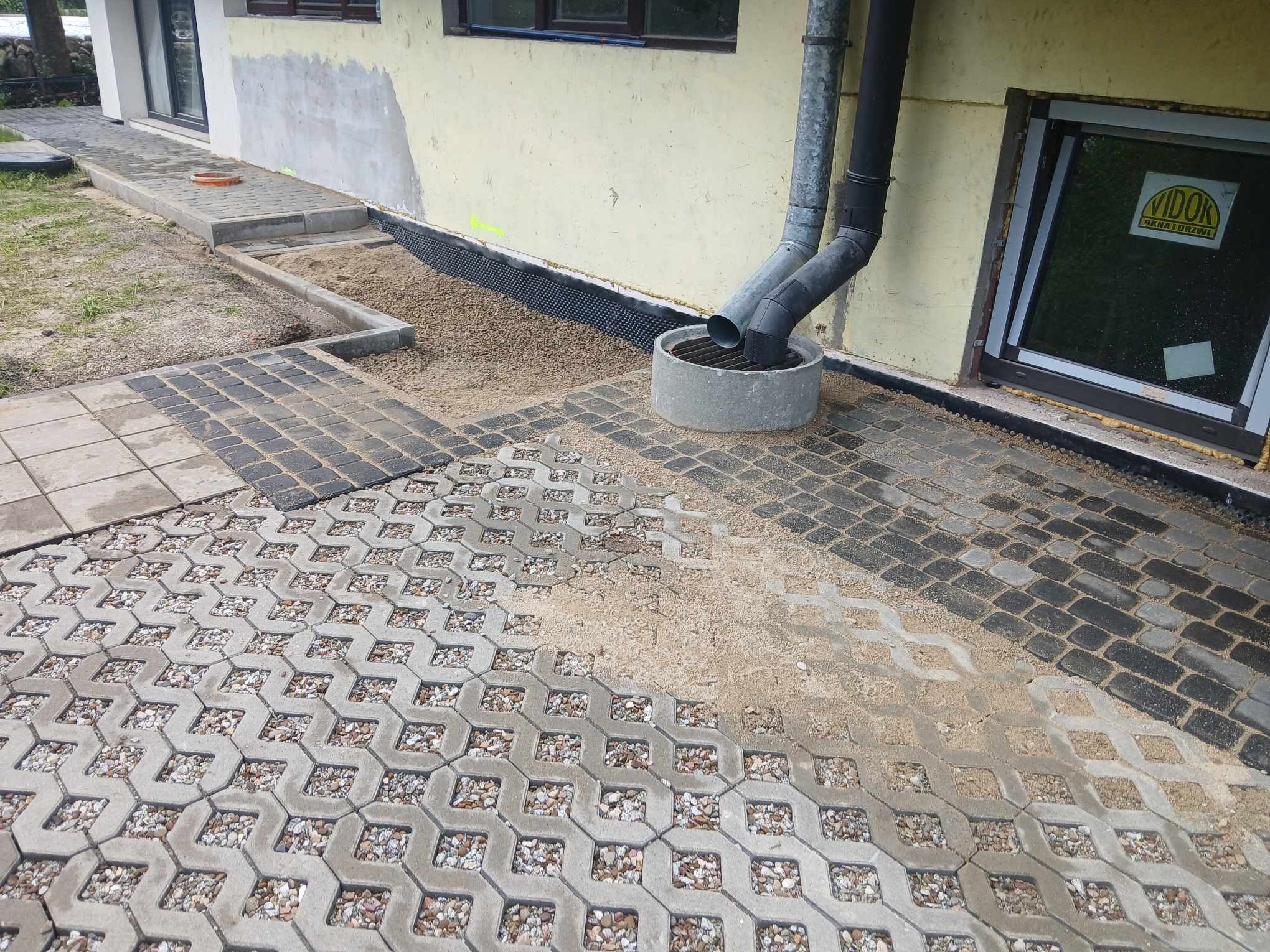 Układanie nawierzchni z ażurowych płyt betonowych wypełnionych kamieniami w Raba Wyżna, widoczny fragment tradycyjnej kostki brukowej i rura spustowa przy budynku.