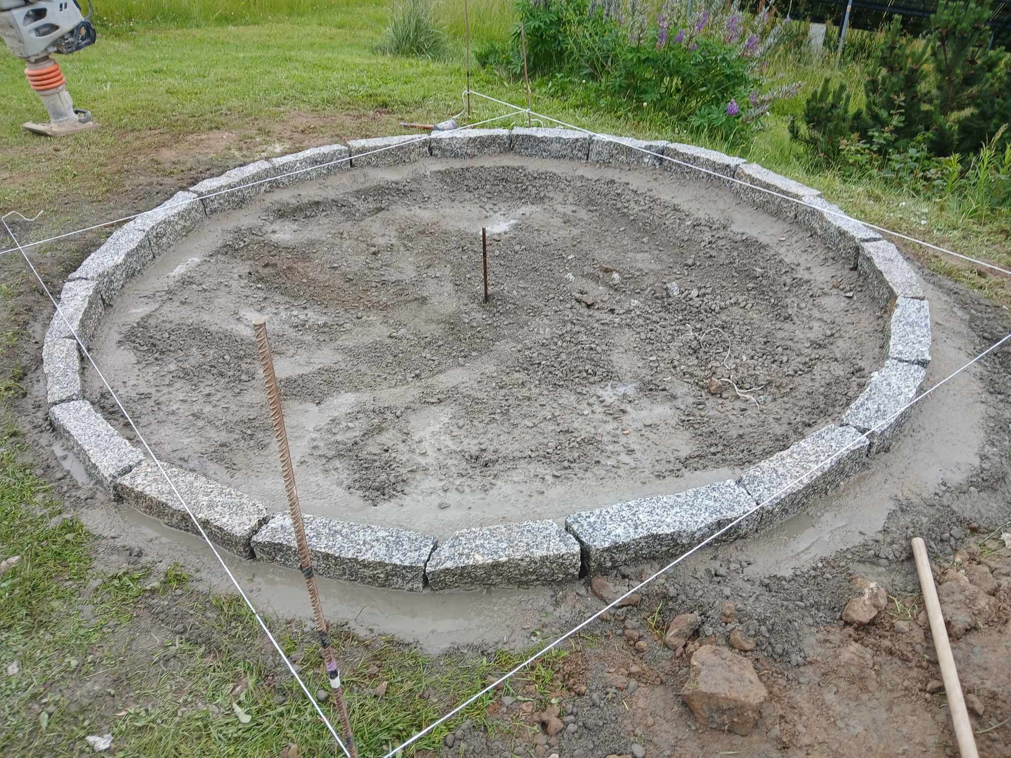 Okrągłe obrzeże z kostki granitowej w trakcie budowy, widoczny ubijak wibracyjny, napięte sznurki wyznaczające promień, podsypka z kruszywa i zaprawy cementowej.