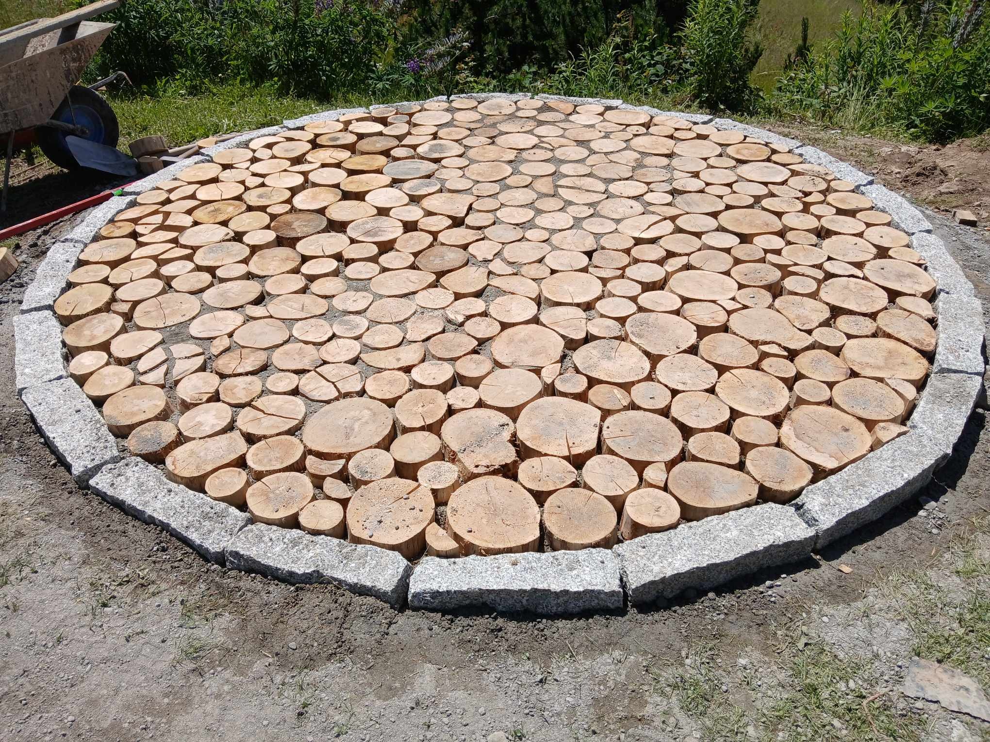 Okrągły plac wyłożony drewnianymi krążkami otoczony szarą kostką granitową, w tle wózek budowlany i zieleń.