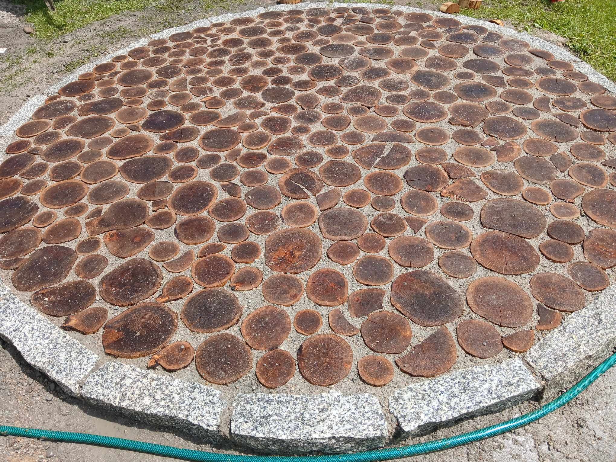 Okrągła ścieżka w ogrodzie wykonana z drewnianych krążków, otoczona szarym obramowaniem z kostki granitowej i zielonym wężem ogrodowym.