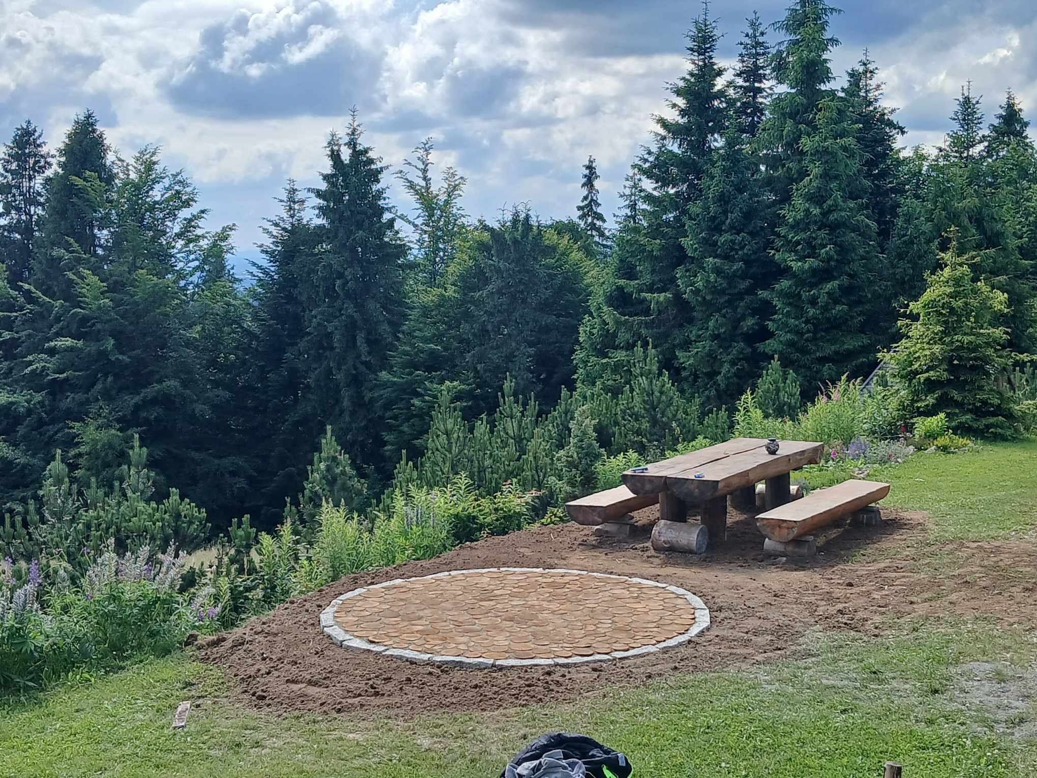 Okrągły plac z drewnianych krążków obramowany jasnoszarą kostką, obok drewniany stół piknikowy z ławkami, w tle las i pochmurne niebo.