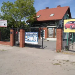 Budynek firmy ATS SECURITY z reklamami systemów zabezpieczeń, w tym automatyki bram i alarmów, widoczny za otwartą bramą z ceglanymi słupkami.