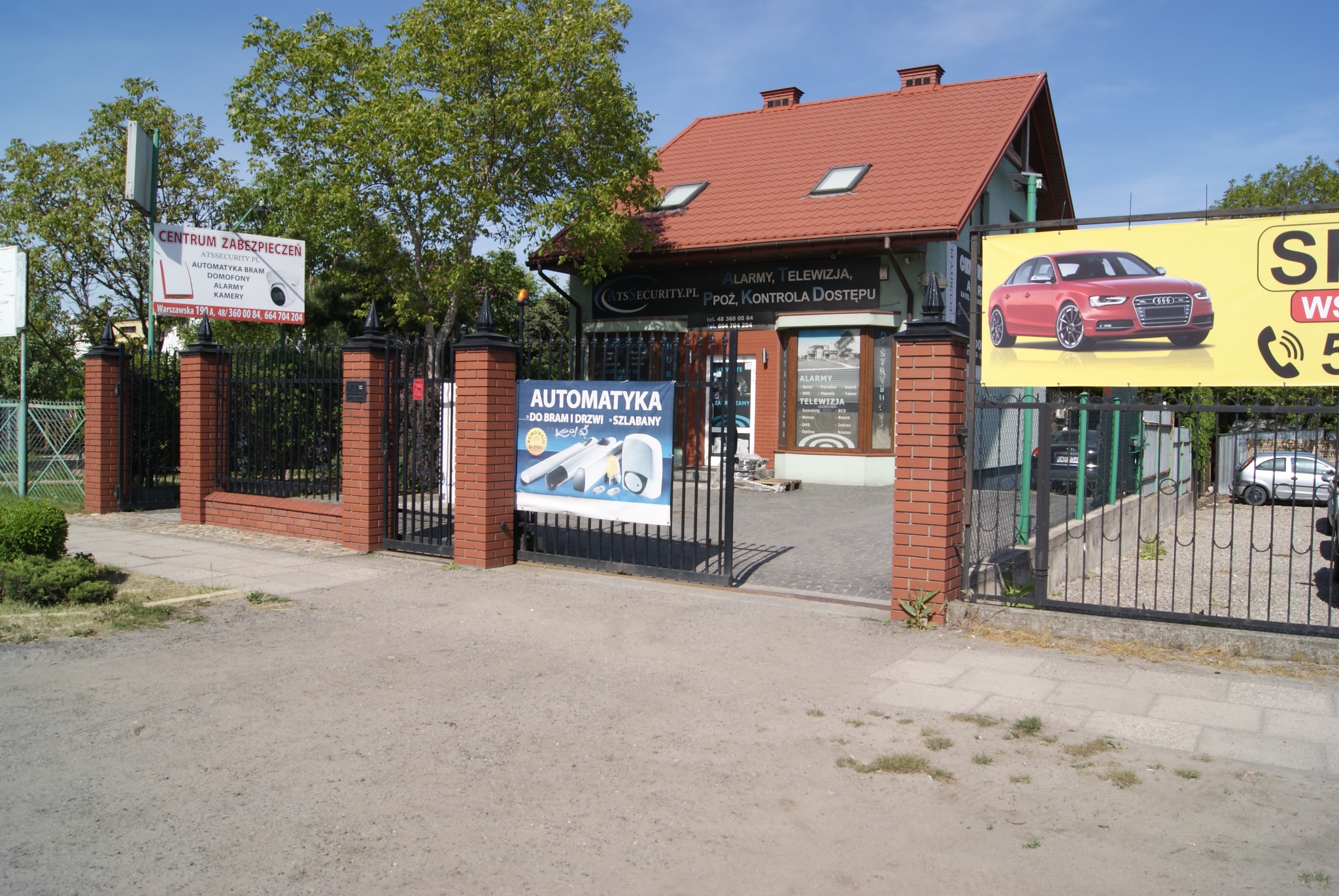 Budynek firmy ATS SECURITY z reklamami systemów zabezpieczeń, w tym automatyki bram i alarmów, widoczny za otwartą bramą z ceglanymi słupkami.
