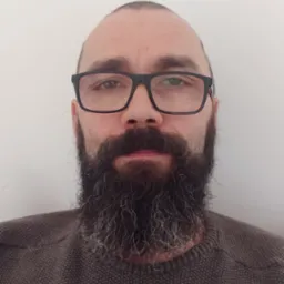 Portret brodatego mężczyzny w okularach na tle białej ściany, ubrany w brązowy sweter o grubym splocie.