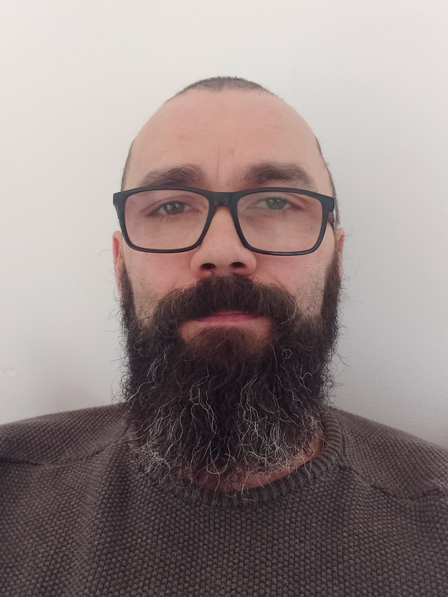 Portret brodatego mężczyzny w okularach na tle białej ściany, ubrany w brązowy sweter o grubym splocie.