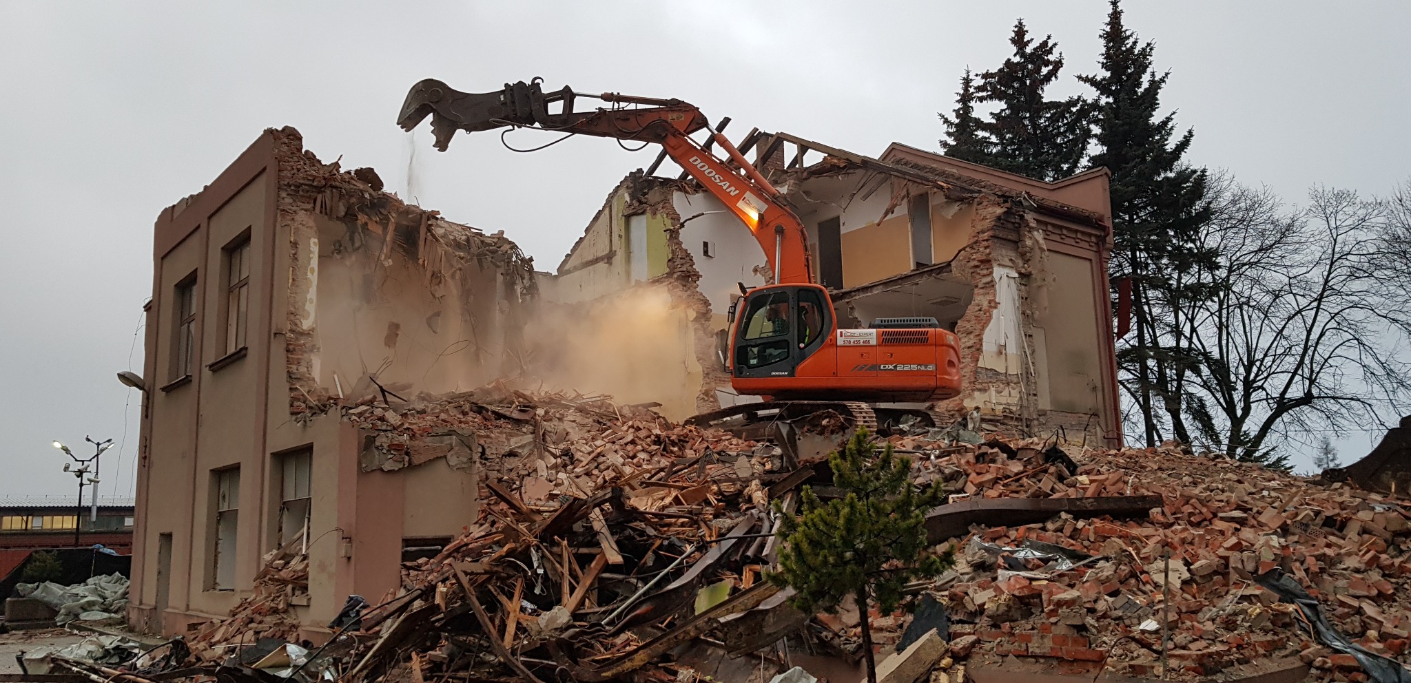 Pomarańczowa koparka Doosan na stercie gruzu, w tle częściowo zburzony budynek z odsłoniętymi ścianami i fragmentami dachu. Praca w toku, widoczny pył.