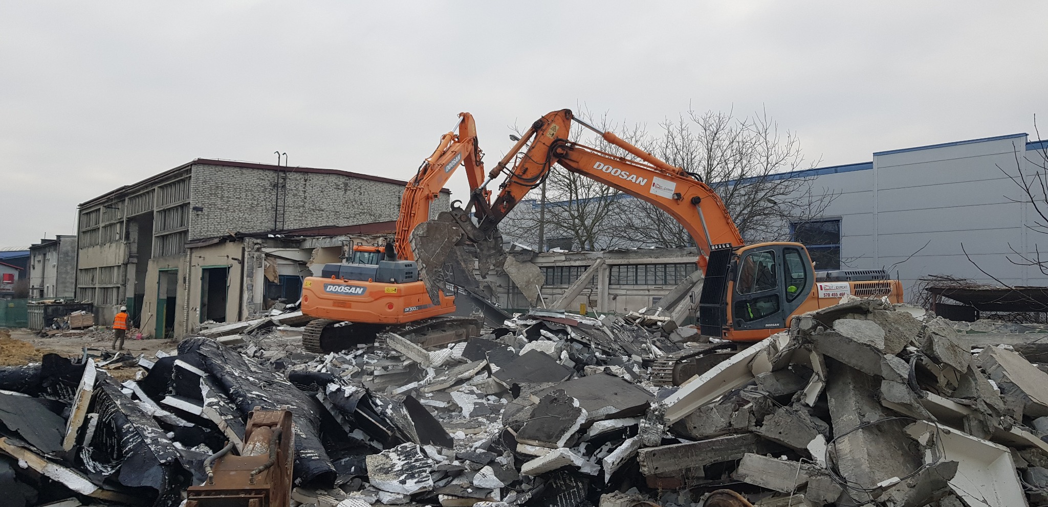 Dwie pomarańczowe koparki Doosan na gruzowisku po rozbiórce budynku. W tle fragmenty zburzonej konstrukcji i pracownik w pomarańczowej kamizelce.