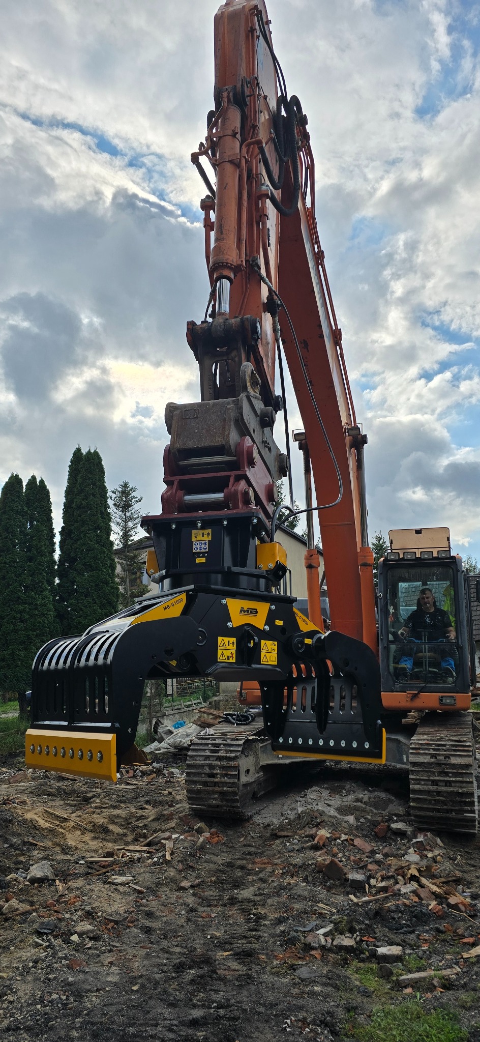 Pomarańczowa koparka z czarno-żółtym osprzętem do kruszenia gruzu, operator w kabinie, teren po rozbiórce, pochmurne niebo w tle.