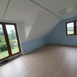 Poddasze po remoncie: świeżo pomalowane ściany na niebiesko i biało, nowe panele podłogowe, okna z widokiem na zieleń. Widoczne puszki elektryczne.