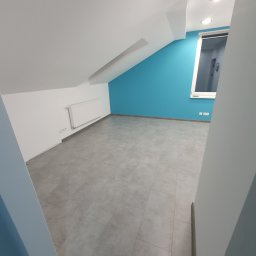 Horizon Amadeusz Glapa - Poddasze po remoncie: błękitna ściana, szara podłoga z płytek, biały grzejnik i okno. Widok z wejścia, skos dachu z lewej strony.