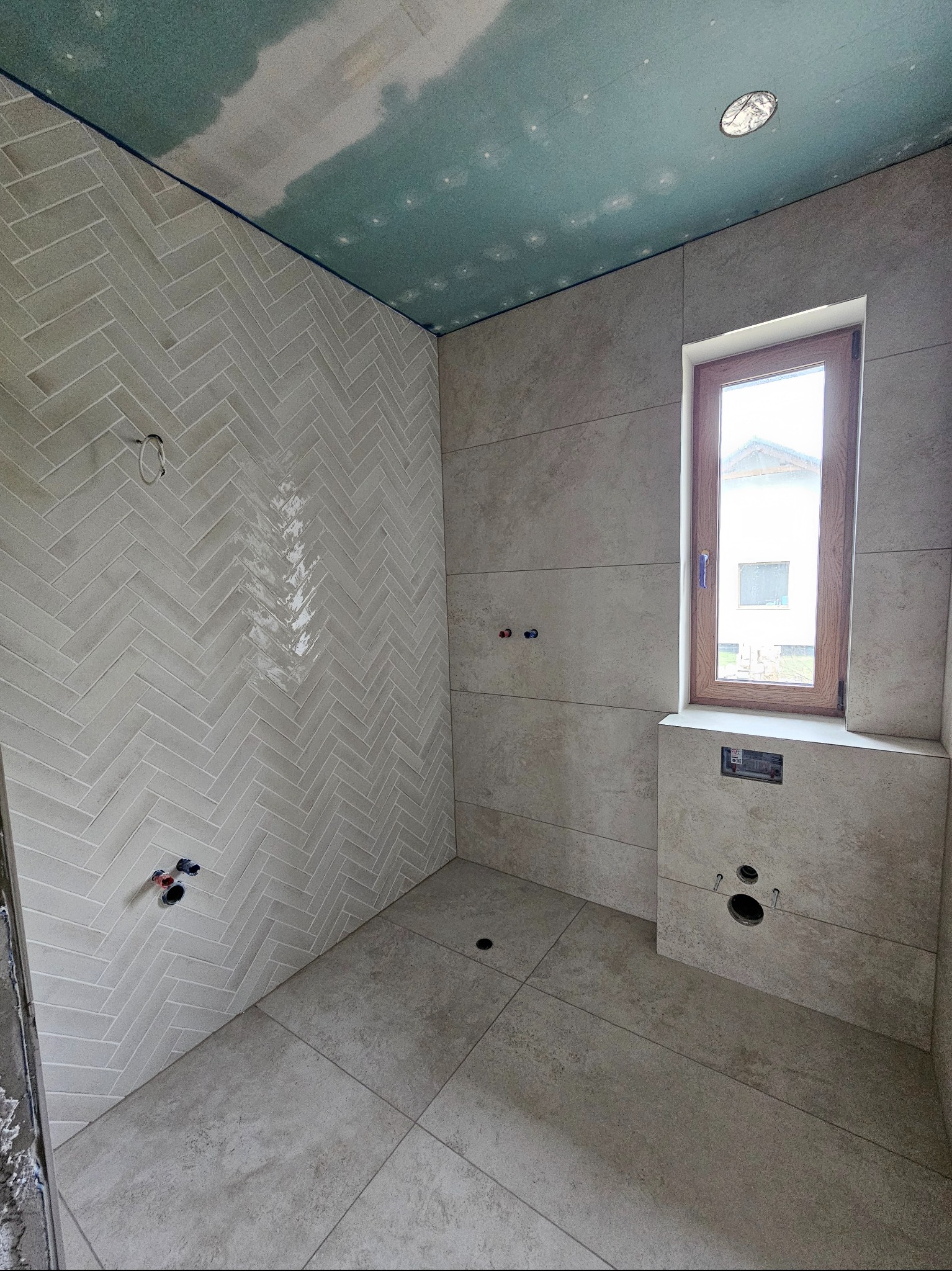 Wykończenie łazienki z beżowymi płytkami ułożonymi w jodełkę na ścianie, widoczne przyłącza wodne, okno i częściowo wykończony sufit z płyt karton-gips.