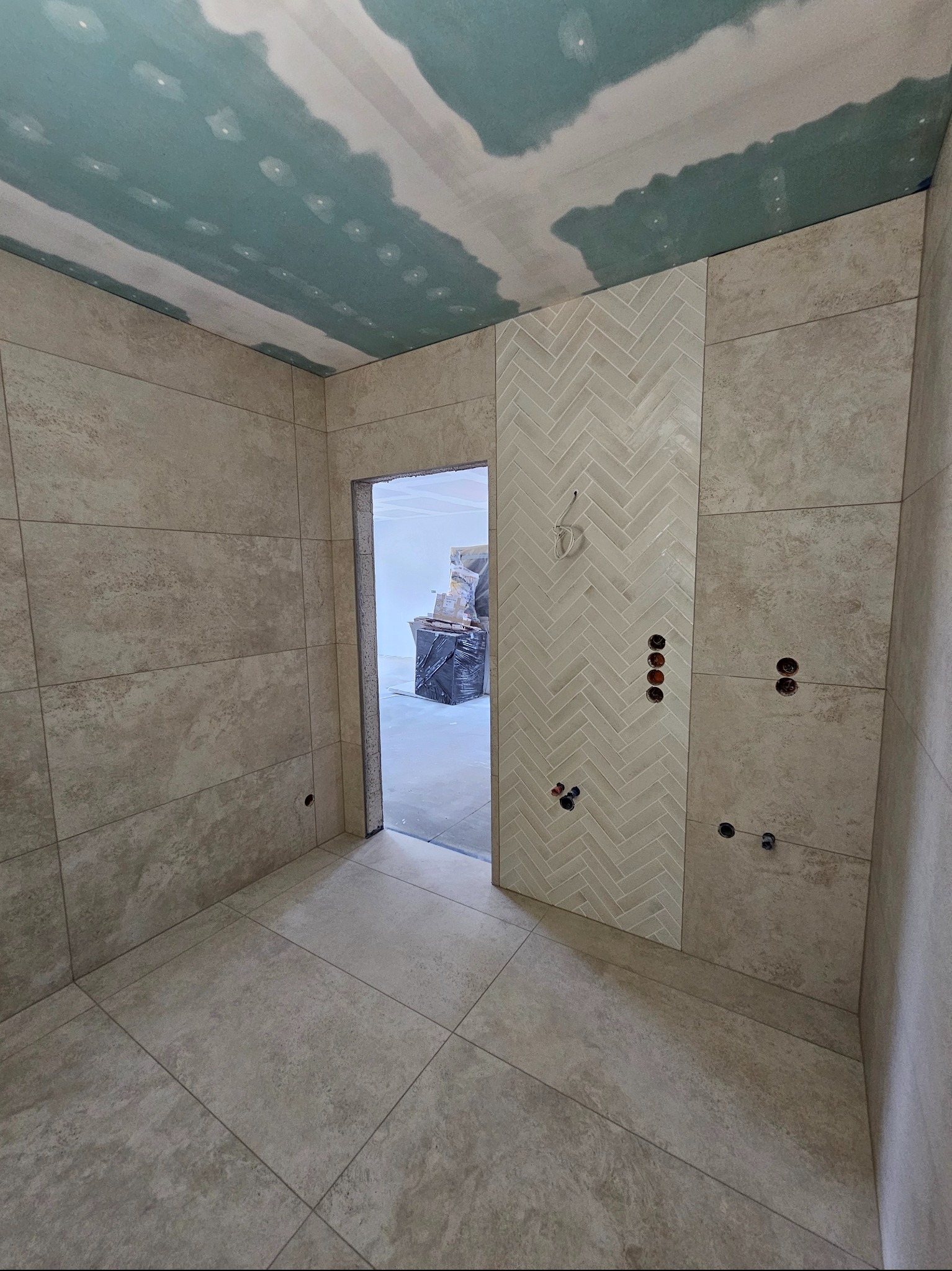 Wykończona łazienka w beżowych płytkach, z dekoracyjnym pasem w jodełkę i przygotowanymi punktami elektrycznymi. Sufit z płyt karton-gips, widok na sąsiednie pomieszczenie.