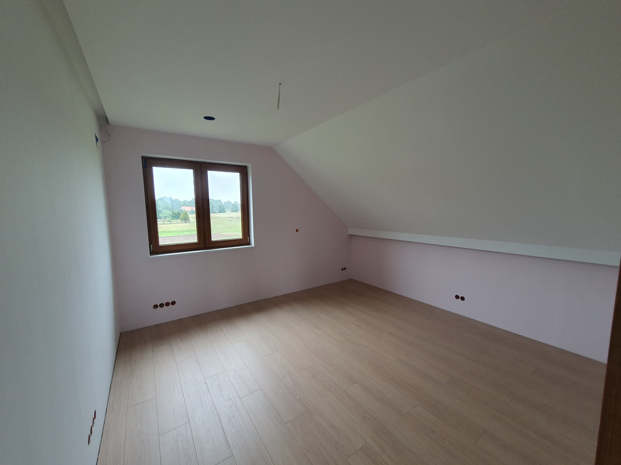 Jasne poddasze po remoncie z oknem, świeżo pomalowanymi ścianami w odcieniach różu i beżową podłogą z paneli. Widoczne gniazdka elektryczne.
