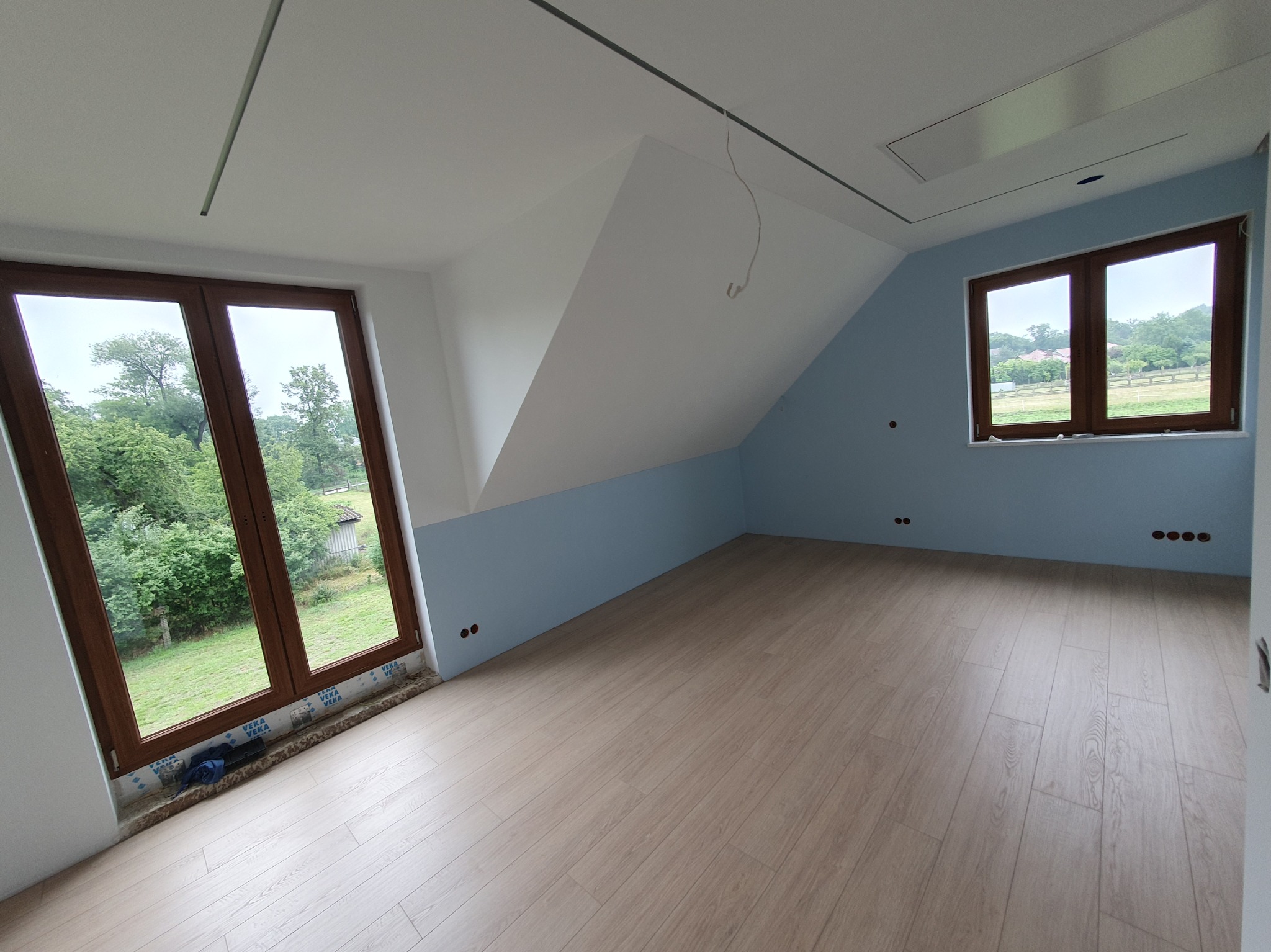 Poddasze po remoncie: świeżo pomalowane ściany na niebiesko i biało, nowe panele podłogowe, okna z widokiem na zieleń. Widoczne puszki elektryczne.
