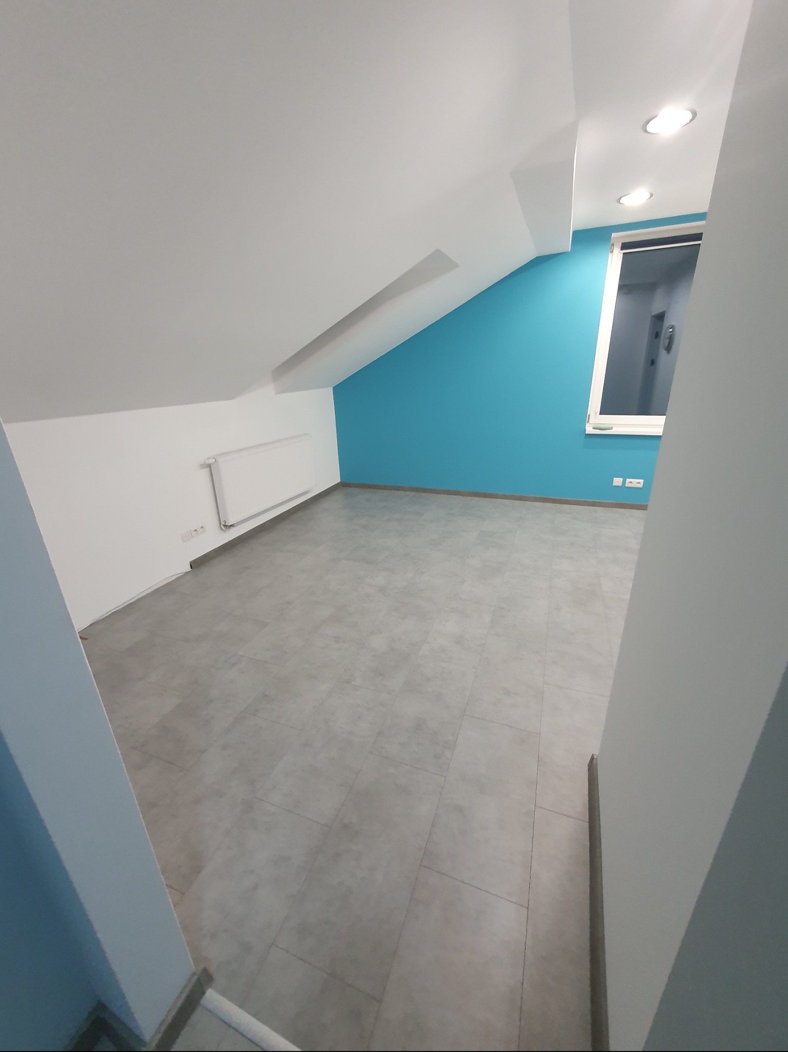 Poddasze po remoncie: błękitna ściana, szara podłoga z płytek, biały grzejnik i okno. Widok z wejścia, skos dachu z lewej strony.