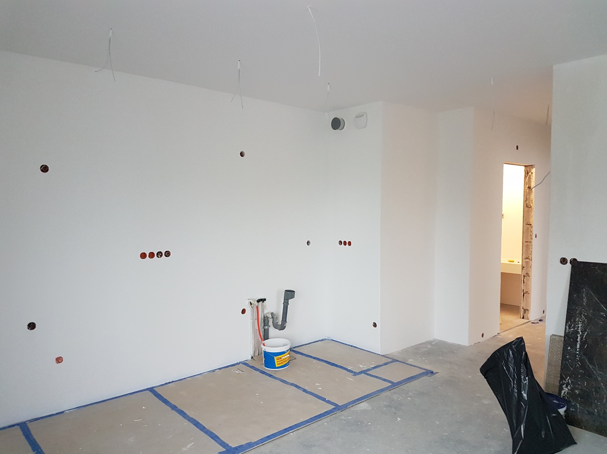 Surowe wnętrze łazienki w trakcie prac wykończeniowych: białe ściany z instalacją, zabezpieczona podłoga, widoczne rury i wiadro farby. Otwór drzwiowy prowadzi do dalszej części łazienki.