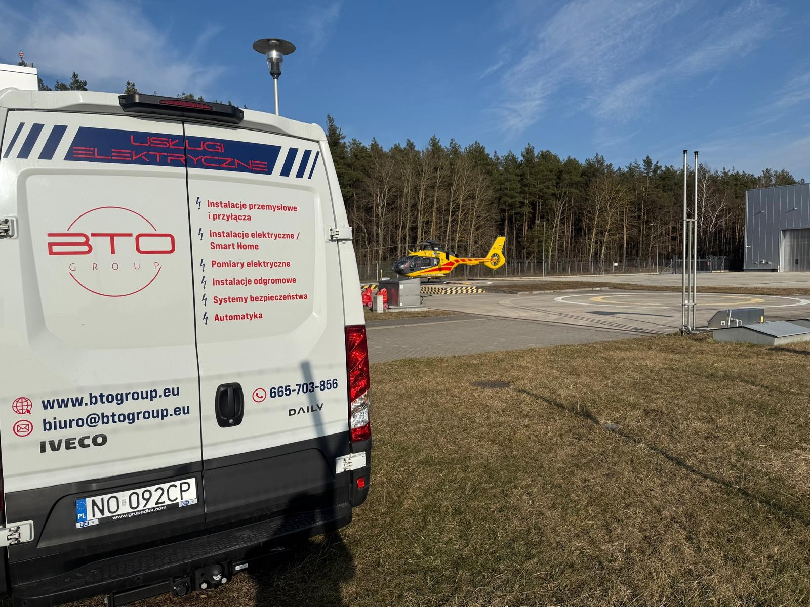 Biały samochód dostawczy z logo BTO Group i ofertą usług: instalacje przemysłowe, elektryczne, pomiary, odgromowe, systemy bezpieczeństwa i automatyka. W tle lądujący helikopter.