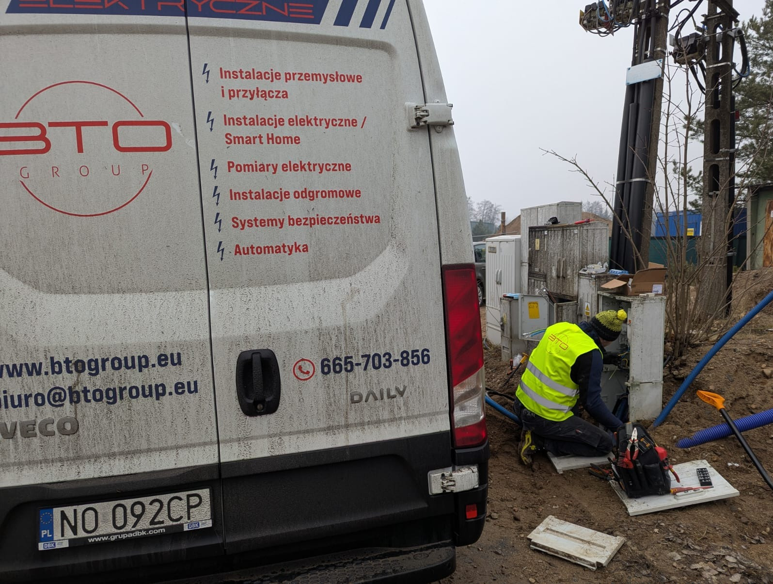 Pracownik w kamizelce BTO Group pracuje przy skrzynce elektrycznej na zewnątrz. W tle zaparkowany brudny bus firmy z wypisanymi usługami: instalacje, pomiary, automatyka.