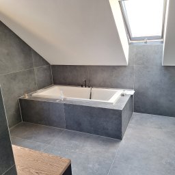 KEMBUD Firma usługowo-budowlana - Nowoczesna łazienka na poddaszu z wanną obudowaną ciemnoszarymi płytkami, podłoga i ściany wyłożone płytkami w podobnym odcieniu, okno dachowe wpuszczające naturalne światło, widoczne akcesoria...