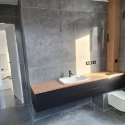 Nowoczesna łazienka z szarymi, wielkoformatowymi płytkami, drewnianym blatem pod umywalką i wiszącą toaletą; widoczne minimalistyczne oświetlenie i otwarte drzwi do innego pomieszczenia.