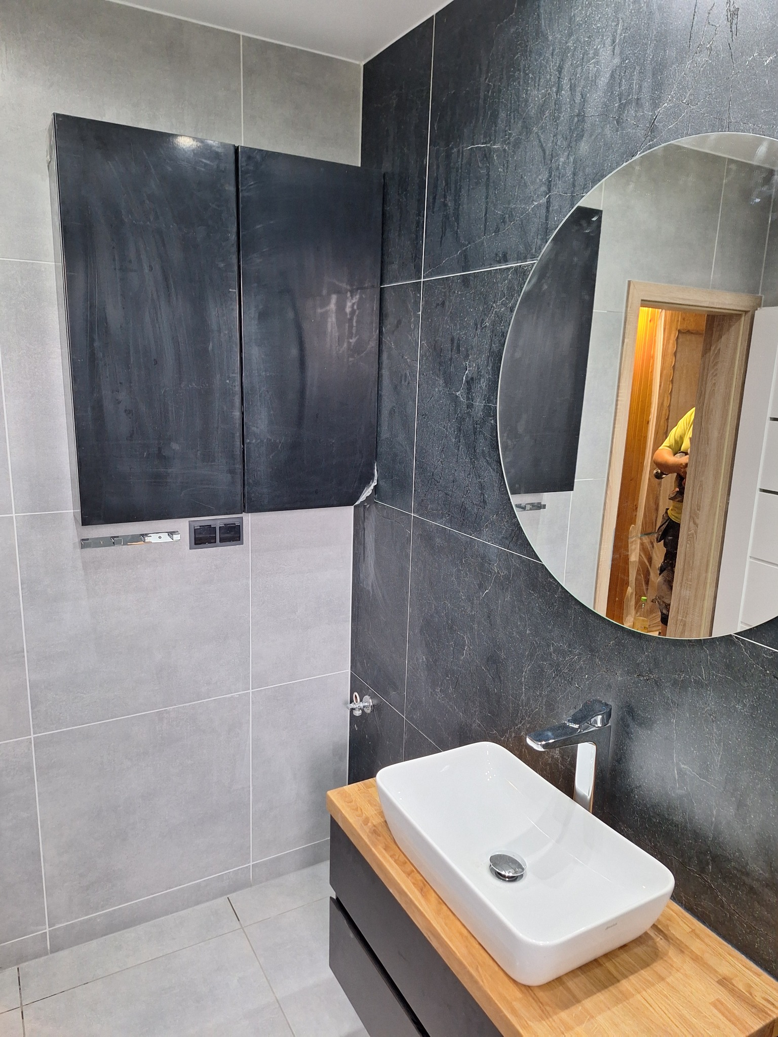 Nowoczesna łazienka z umywalką nablatową na drewnianym blacie, czarnymi szafkami wiszącymi i lustrem, w którym odbija się osoba w żółtej koszulce podczas prac wykończeniowych.