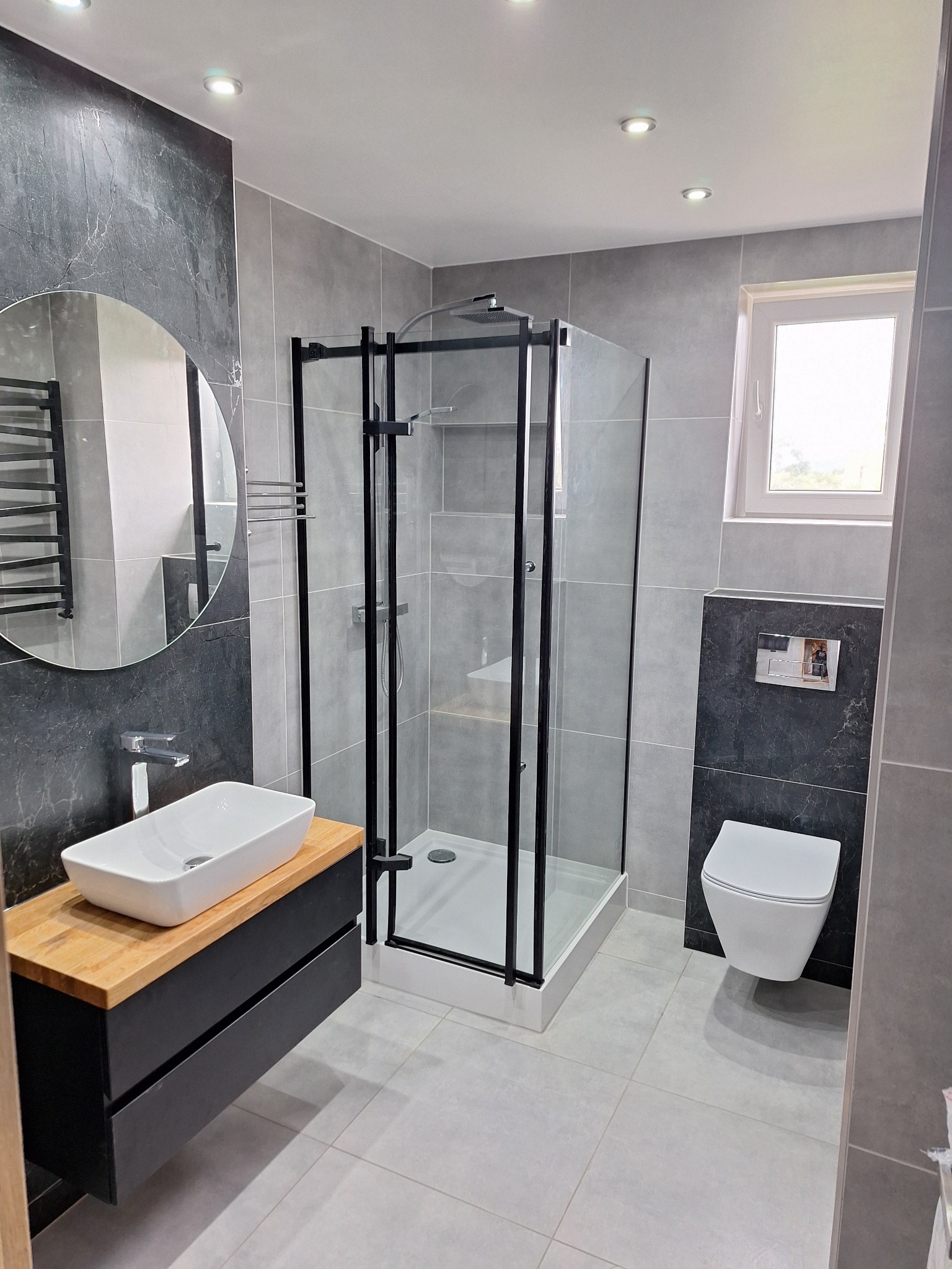 Nowoczesna łazienka z szarymi płytkami, kabiną prysznicową z czarną ramą, wiszącą toaletą i umywalką nablatową na drewnianej szafce.