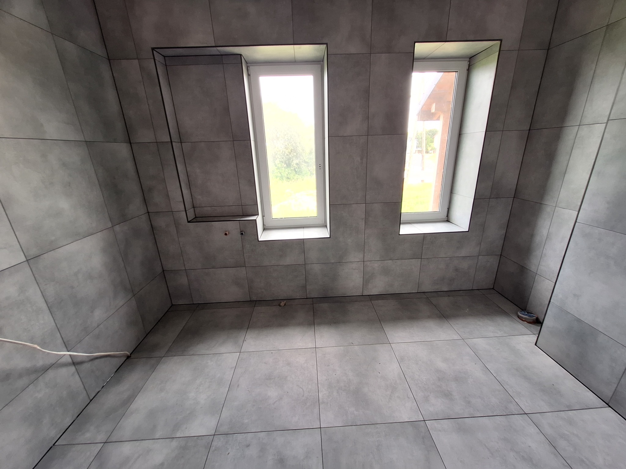 Wykończona łazienka w szarościach z dwoma oknami i wnęką, widoczne płytki na ścianach i podłodze, kabel elektryczny wystający z podłogi.