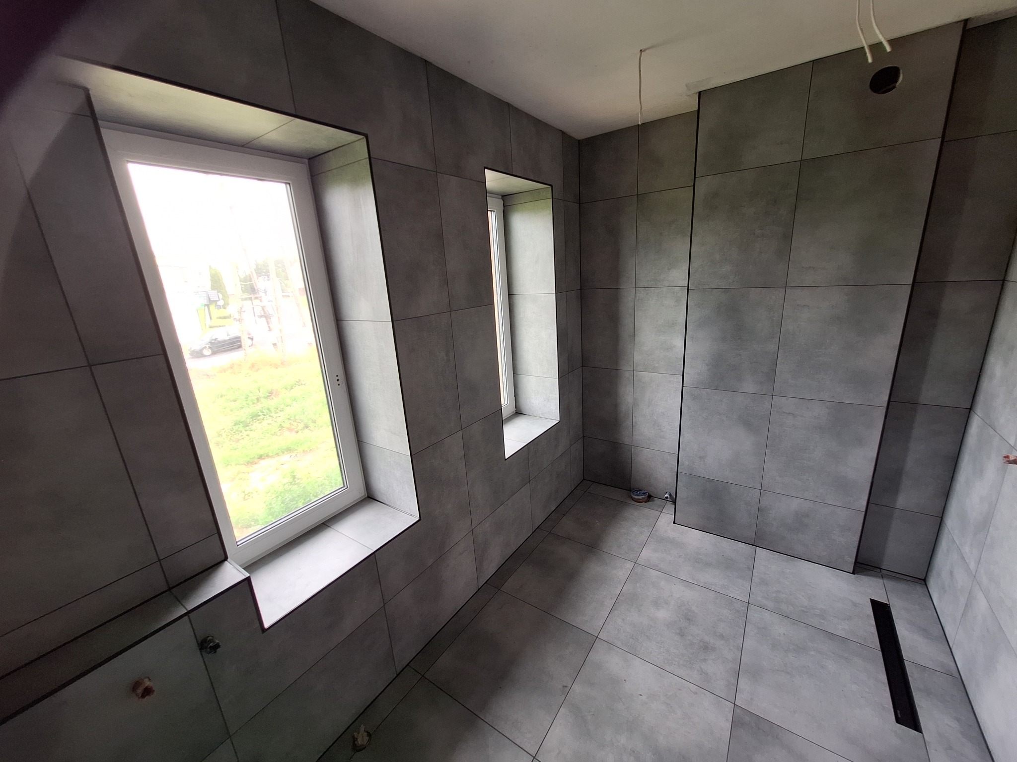 Wykończona łazienka z szarymi, kwadratowymi płytkami na ścianach i podłodze, dwa okna z białymi ramami wpuszczone w ścianę, widoczny odpływ liniowy w podłodze.