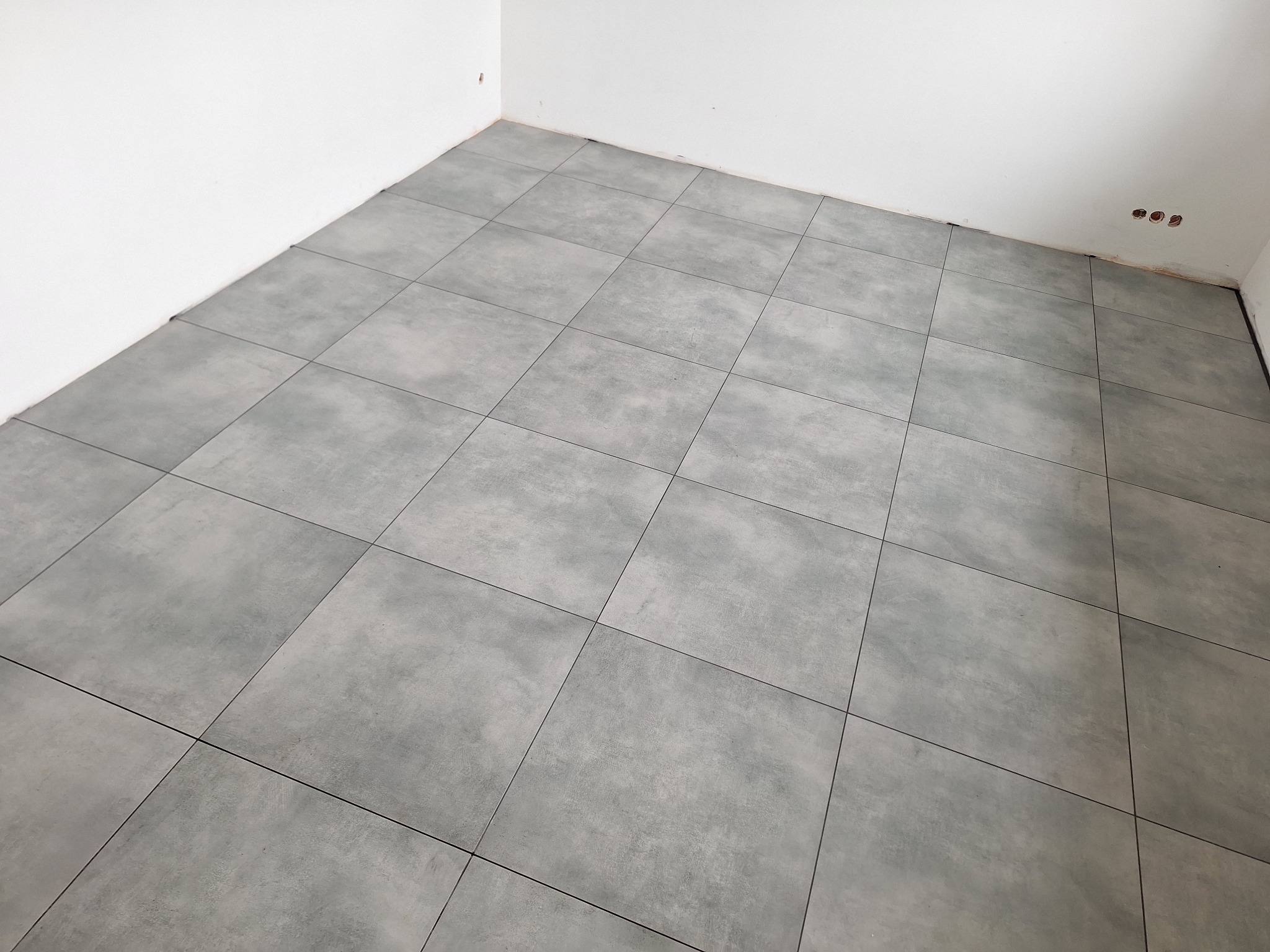 Ułożone szare płytki podłogowe w rogu pokoju z białymi ścianami, widoczne trzy otwory na gniazdka elektryczne w ścianie.