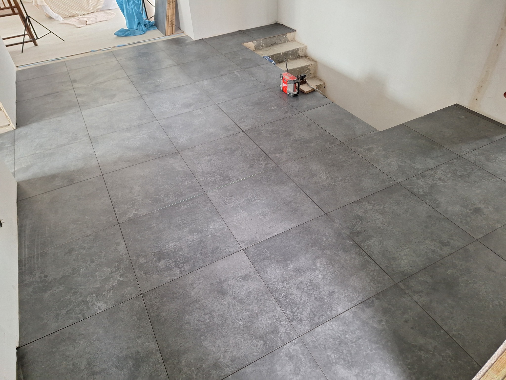 Ułożone szare płytki podłogowe o matowym wykończeniu, z widocznym poziomnicą laserową w pobliżu schodów w trakcie remontu.
