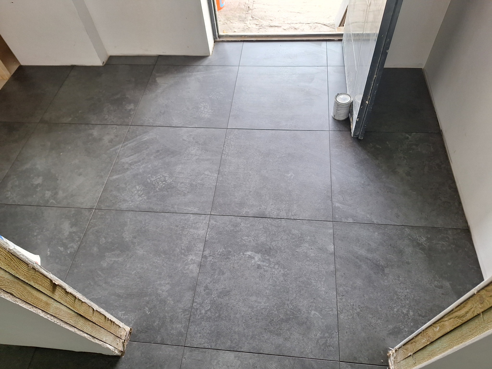 Ułożone szare płytki podłogowe w korytarzu, widoczna puszka z farbą przy drzwiach i fragmenty ościeżnic.