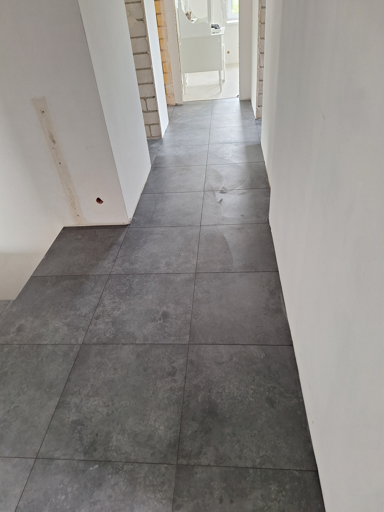 Ułożona, szara, matowa podłoga z płytek w korytarzu, widoczne ślady po remoncie na ścianach i surowy stan wykończenia w przejściu do kolejnego pomieszczenia.