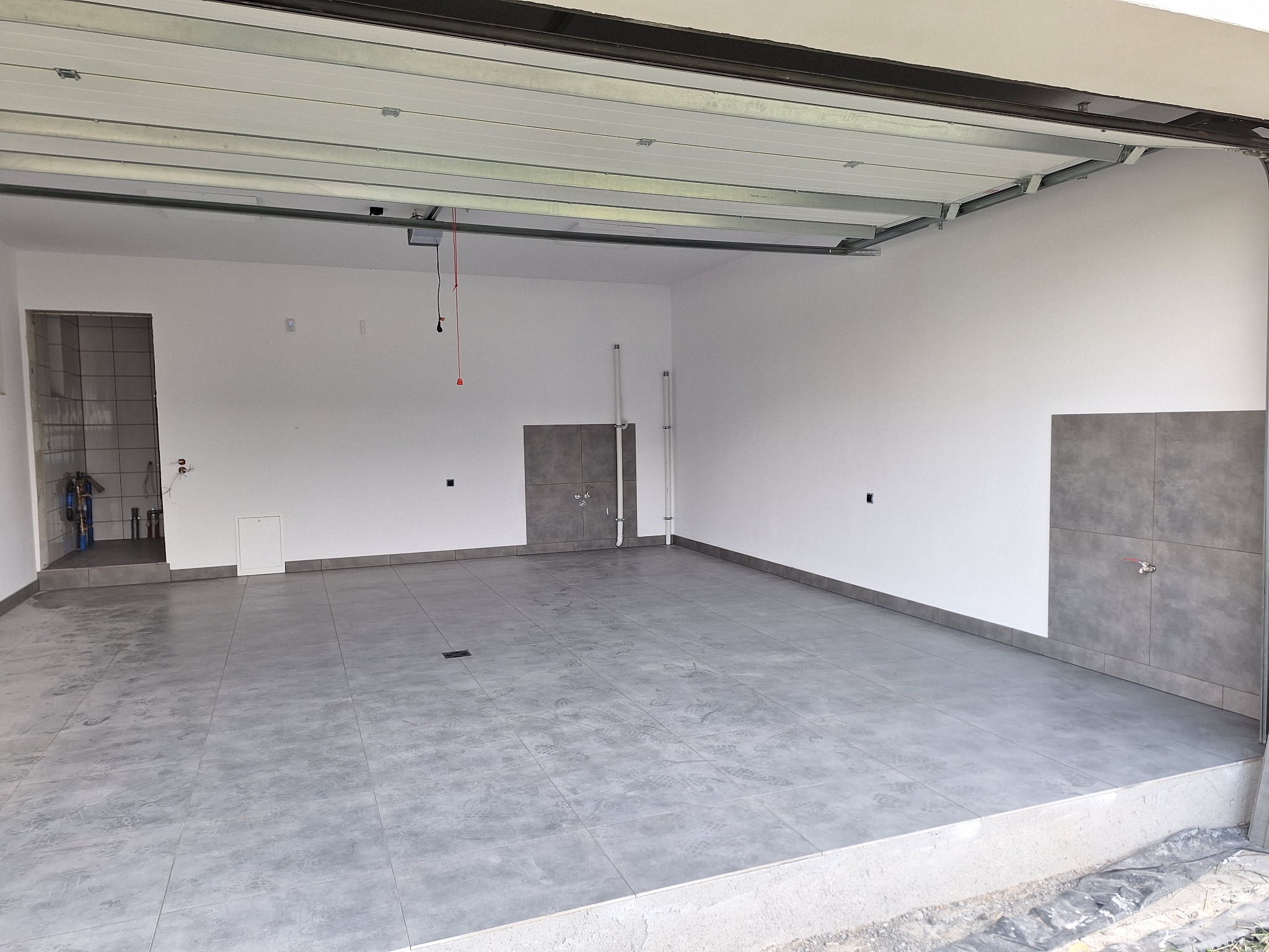 Wnętrze garażu po remoncie z szarą podłogą z płytek, fragment ściany z płytkami i instalacją wodną, widoczny otwarty wjazd z bramą garażową.