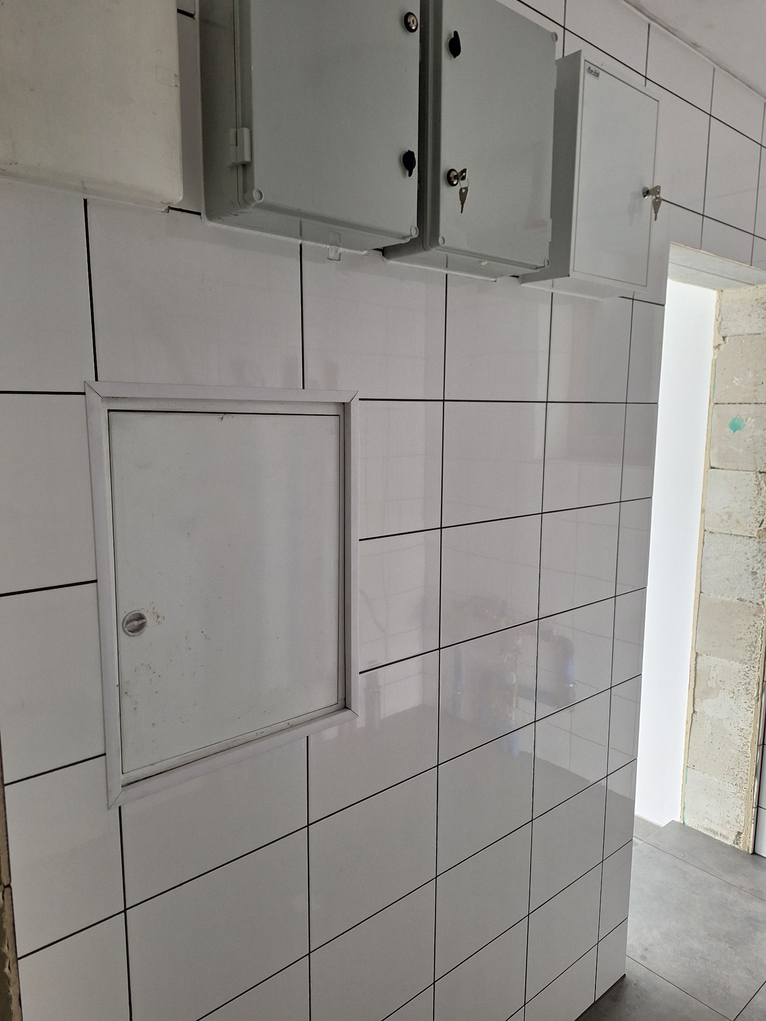Białe, błyszczące płytki ceramiczne na ścianie z widocznymi trzema szafkami elektrycznymi z kluczami i inspekcyjnym włazem rewizyjnym.