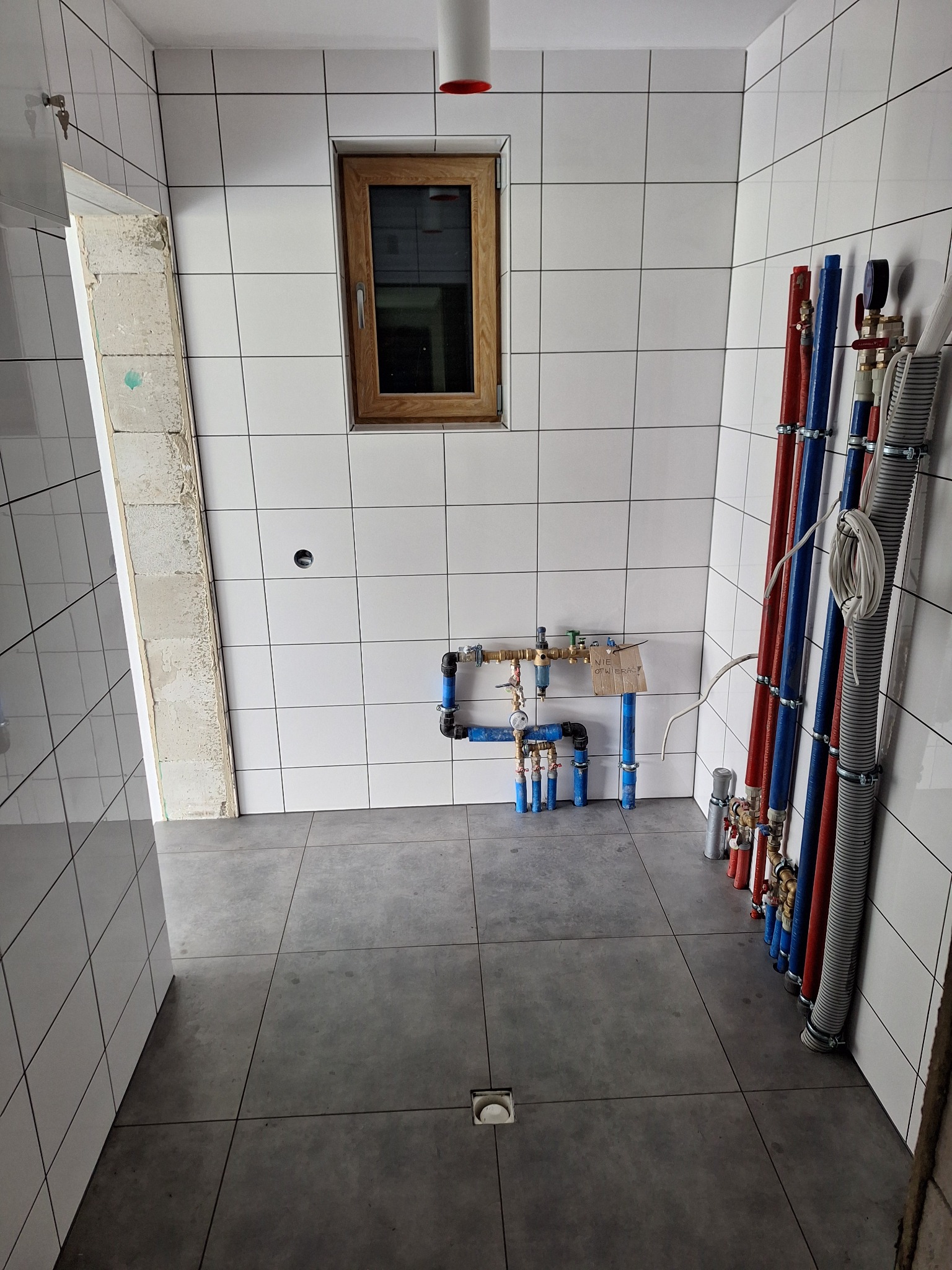 Wnętrze łazienki w trakcie remontu: białe płytki ścienne z czarną fugą, szara podłoga z odpływem liniowym, widoczne rury instalacyjne w kolorach czerwonym i niebieskim, okno z drewnianą ramą...