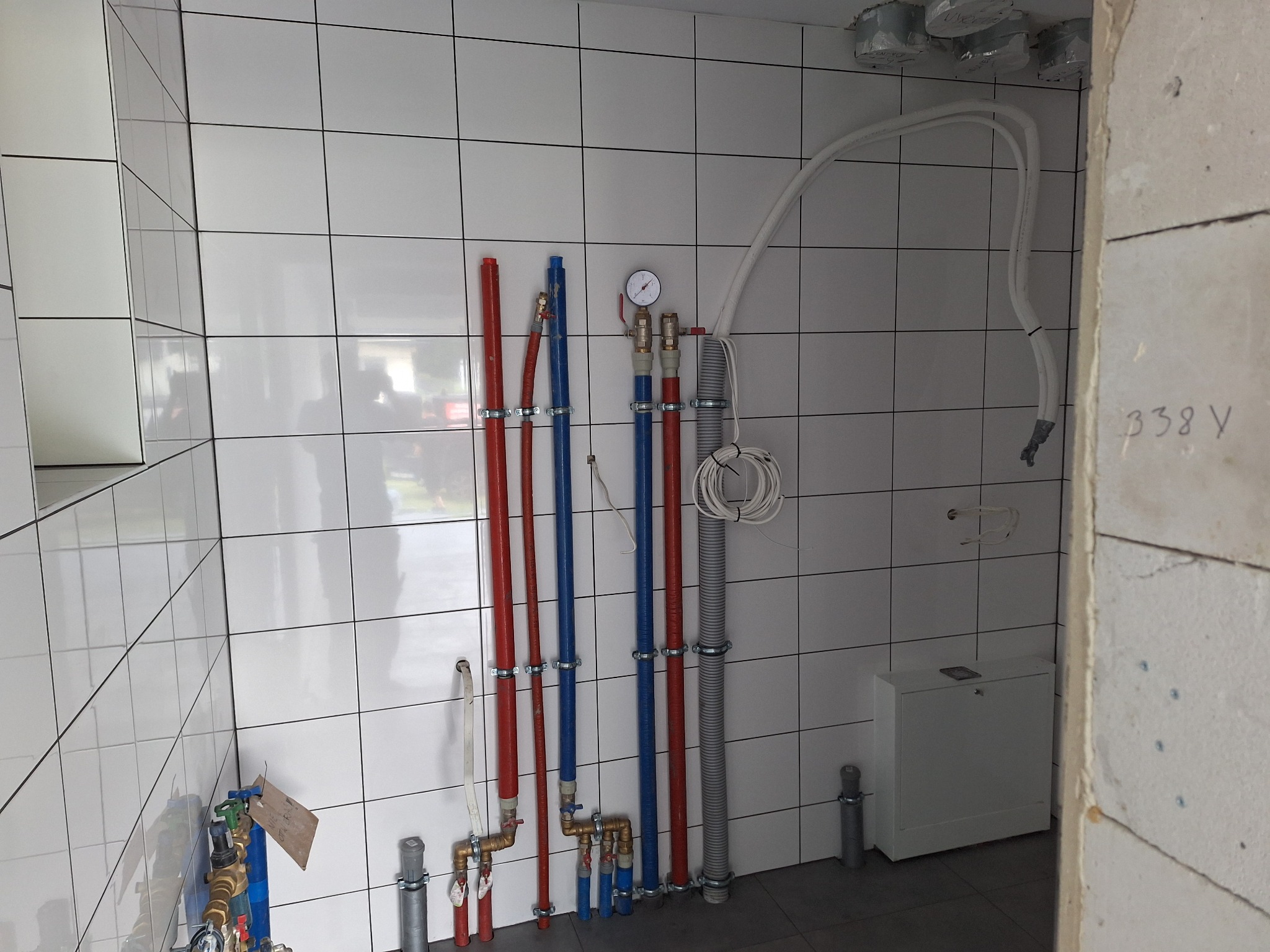 Świeżo ułożone, białe, kwadratowe płytki na ścianie w trakcie remontu łazienki, widoczne rury wodne w kolorach czerwonym i niebieskim, zamontowane na metalowych uchwytach, oraz manometr.