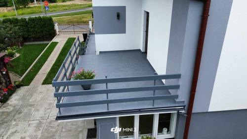 Szary balkon z drewnianą balustradą i donicą z różowymi kwiatami, widok z góry, na zewnątrz widoczny zadbany ogród i droga.