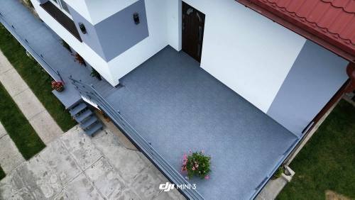 Widok z góry na nowoczesny balkon z szarą, gładką powierzchnią, biało-szarą elewacją budynku, metalową barierką i doniczką z kwitnącymi różowymi kwiatami w rogu.