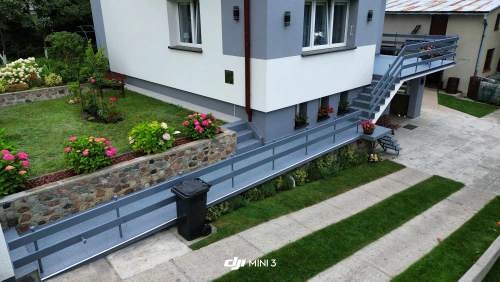 Widok z góry na odnowioną elewację budynku z szarymi akcentami, schody z balustradą prowadzące na balkon, kamienne murki oddzielające rabaty kwiatowe oraz trawnik z pasami zieleni.