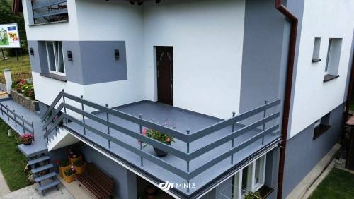 Dwukondygnacyjny dom z balkonem i tarasem, pomalowany na biało-szaro, z szarą balustradą, kwiatami w donicach i widocznymi schodami wejściowymi.