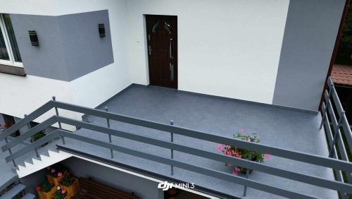 Wykończony balkon z szarą podłogą i barierkami, widok z góry z lekkim ujęciem na schody, kwiaty w donicach i elewację budynku z zewnętrznymi lampami.