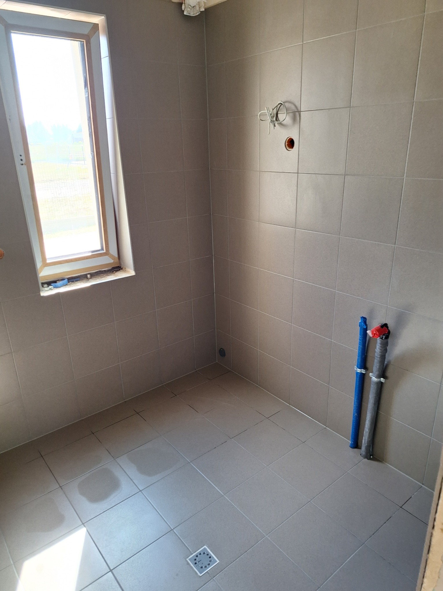 Wnętrze łazienki w trakcie prac wykończeniowych, widoczne szare płytki na ścianach i podłodze, okno z widokiem na zewnątrz oraz rury przy ścianie.