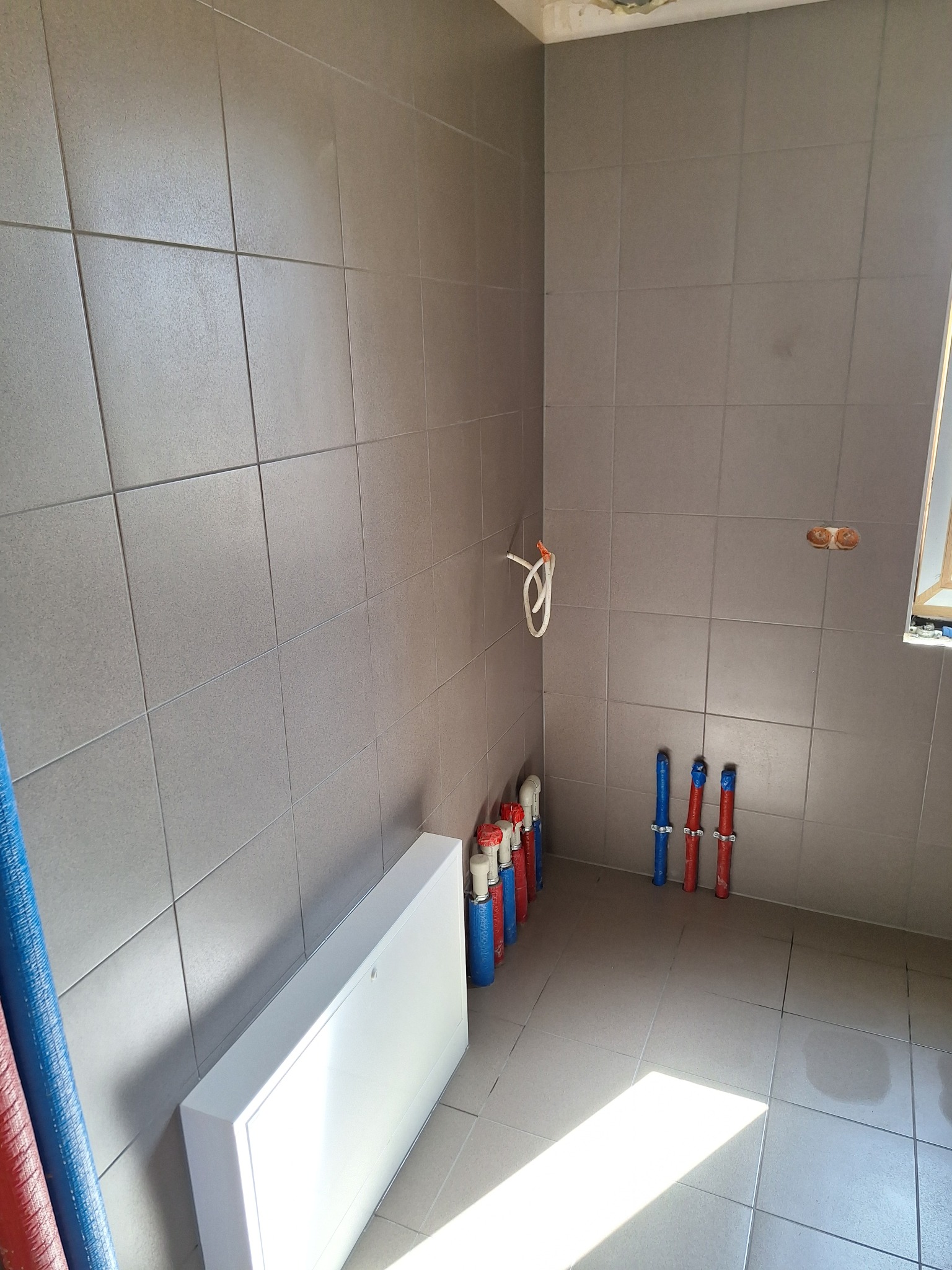 Ściany łazienki wyłożone szarymi płytkami, widoczne rury instalacyjne, biała obudowa rewizyjna i częściowo okno. Niedokończone prace wykończeniowe.